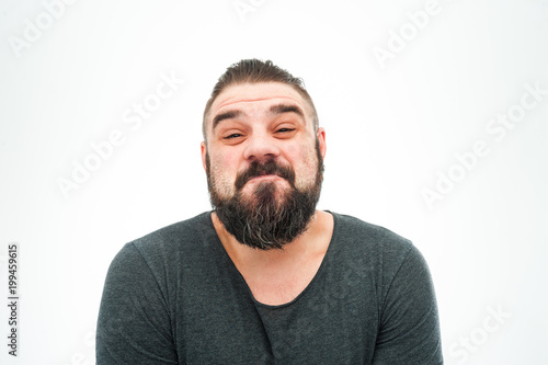 Good looking cute young bearded male looks with hesitant expression as doubts what to choose, points with thumb aside, copy space for promotional text or advertisment