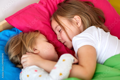 childhood, bedtime and family concept - happy little girls or sisters sleeping in bed at home