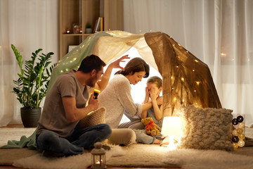 hygge and people concept - father with torch light telling scary stories to his daughter and wife, family having fun in kids tent at night at home