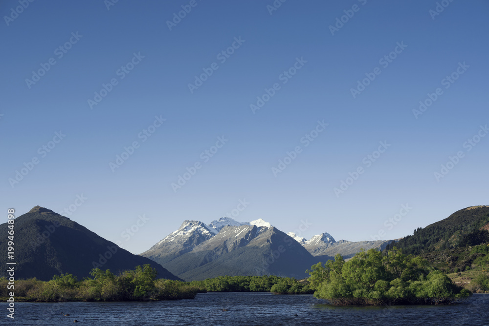 Paisaje de montañas con picos nevados frente a un gran lago de agua muy azul al atardecer. Cielo azul despejado.