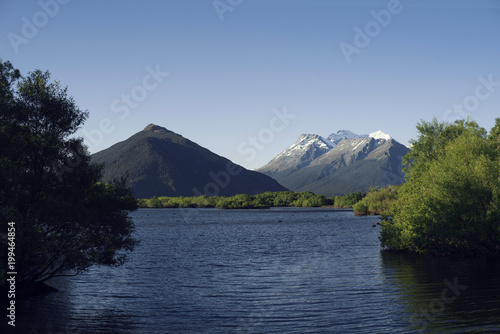 Paisaje de montañas con picos nevados frente a un gran lago de agua muy azul al atardecer. Hay árboles en primer plano. Cielo azul despejado.