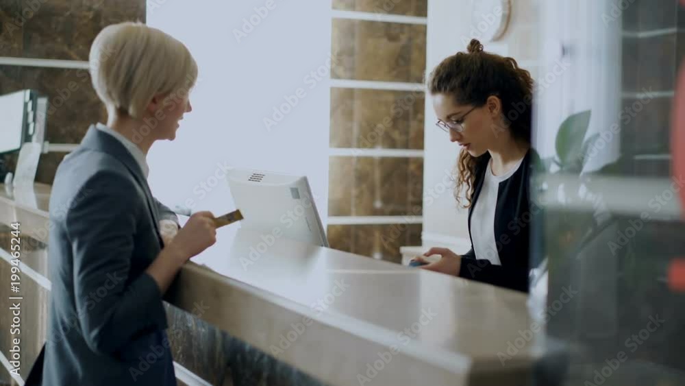 Businesswoman guest in hotel reception paying bill contactless payment credit card technology and talking with receptionist girl standing at desk. Business, travel and people concept Businesswoman guest in hotel reception paying bill contactless payment credit card technology and talking with receptionist girl standing at desk. Business, travel and people concept