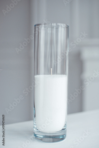 A burning candle in tall glass vase flask standing on the table indoor.