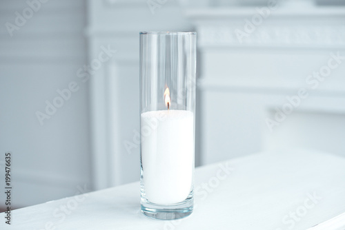 A burning candle in tall glass vase flask standing on the table indoor.