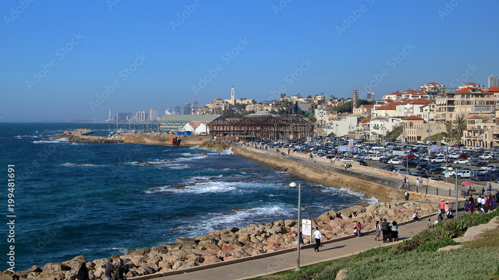 Widok na port w Jafie, Izrael, ze wzgórza, wody Morza Śródziemnego ze spienionymi falami, umocnienia na brzegu, ludzie spacerujący po promenadzie nadmorskiej, w dali nowoczesna zabudowa Tel Awiwu 