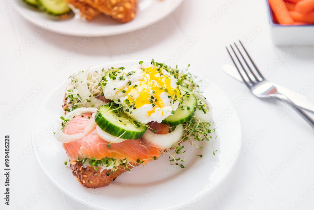 Cereal bun with egg benedict, smoked salmon, sprout micro greens, onion and cucumber slices, cream cheese on the served white wooden table. Healthy breakfast concept. Selective focus.