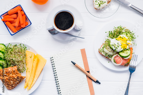Top view planning notebook with copy space and healthy breakfast. Sandwich with egg benedict and smoked salmon, plate with cheese, sprout micro greens and cucumber, carrots and coffee on wooden table. photo