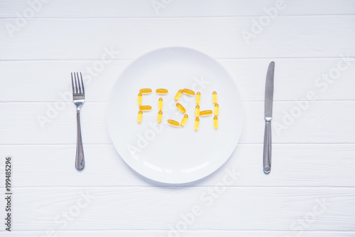 Top view white plate with Fish word lettering by cod Liver Oil Capsules, Omega 3 served with knife and fork on the white wooden table. Healthy living concept. Selective focus, space for text.