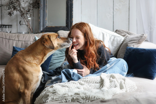 Hübsche rothaarige Frau sitzt auf einem Sofa lacht und wird von einem Hund angestupst