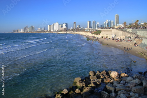 Zatoka Morza Śródziemnego w Tel Awiwie, niebieska czysta woda, kamienista i piaszczysta plaża z turystami, na horyzoncie wyoskie, jane, nowoczesne budynki miasta, bezchmurne błękitne niebo