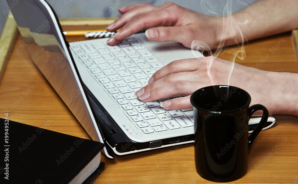 Women's fingers on the keyboard of the laptop that is on the table with a book and cup of coffee Women's fingers on the keyboard of the laptop that is on the table with a book and cup of coffee