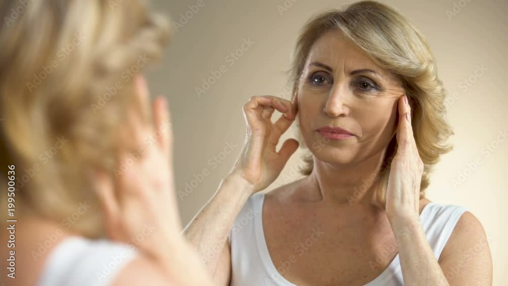 Unhappy aged woman looking in mirror at home, touching her face, aging process Unhappy aged woman looking in mirror at home, touching her face, aging process