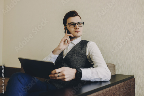 Young businessman sitting on a sofa with a computer on kness and talking on phone. photo