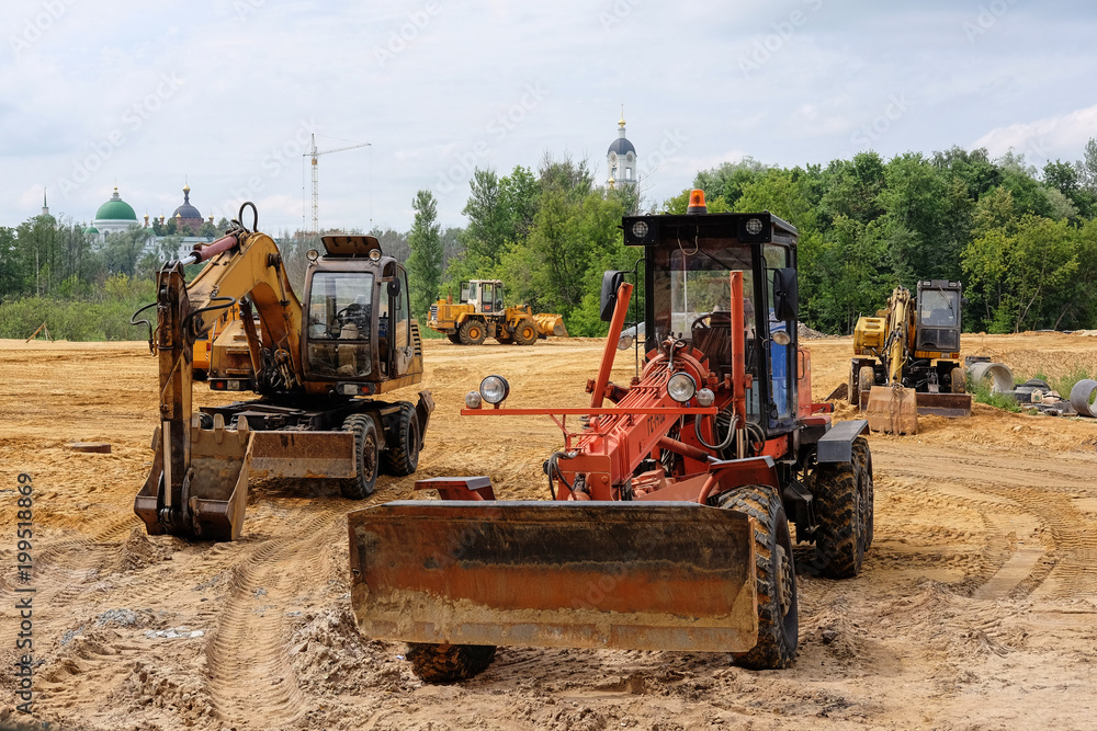 excavators and bulldozers on a large sandy field, behind which there is a forest, an hour tower and a church excavators and bulldozers on a large sandy field, behind which there is a forest, an hour tower and a church