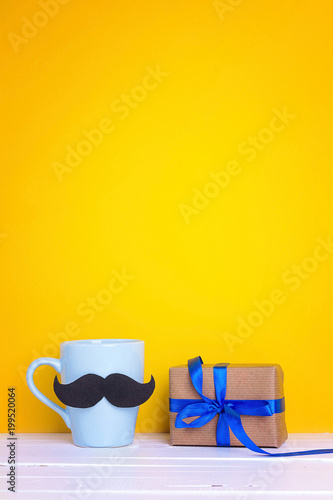 Blue mug with a mustache and gift box on yellow background. Copy space. Fathers day holiday.