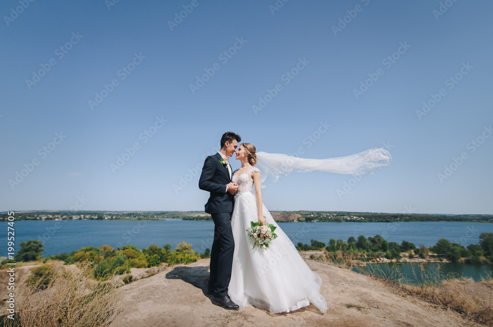 Beautiful bride and groom in a lace dress and flying veil stand against the background of cliffs and the sea. Solar wedding of the newlyweds. Beautiful bride and groom in a lace dress and flying veil stand against the background of cliffs and the sea. Solar wedding of the newlyweds.