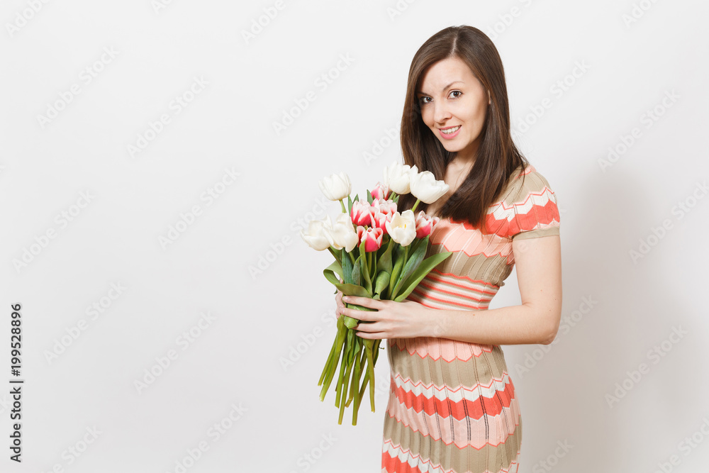 Beautiful young smiling brunette girl in light patterned dress is holding bouquet of white and pink tulips in hands, smiling and rejoices in studio on white background. Concept of holiday, good mood. Beautiful young smiling brunette girl in light patterned dress is holding bouquet of white and pink tulips in hands, smiling and rejoices in studio on white background. Concept of holiday, good mood.