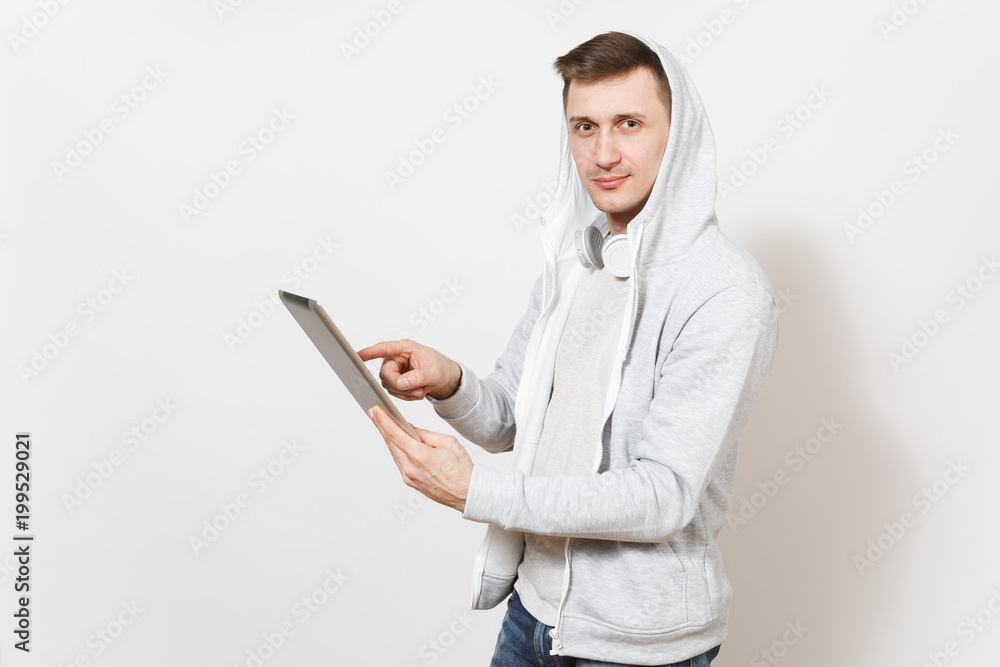 Young handsome man tudent in t-shirt and light sweatshirt with hood with headphones holds tablet pc computer in hands, using it and looks at camera isolated on white background. Concept of technology Young handsome man tudent in t-shirt and light sweatshirt with hood with headphones holds tablet pc computer in hands, using it and looks at camera isolated on white background. Concept of technology