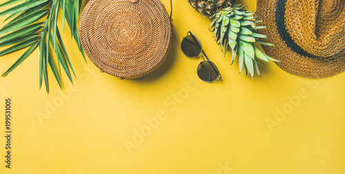 Colorful summer female fashion outfit flat-lay. Straw hat, bamboo bag, sunglasses, palm branches, pineapple over yellow background, top view, copy space, wide composition. Summer fashion, holiday
