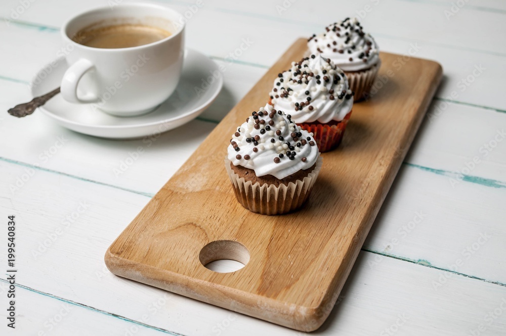 Vanilla cupcakes with chocolate topping and cup of coffee on a wooden chopping board on a light background Vanilla cupcakes with chocolate topping and cup of coffee on a wooden chopping board on a light background