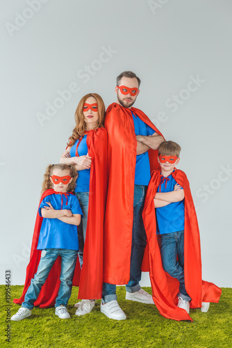 family in superhero costumes standing with crossed arms and looking at camera on grey