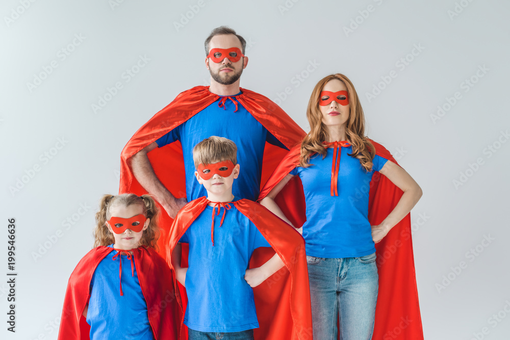 family of superheroes in costumes standing with hands on waist and looking at camera isolated on grey