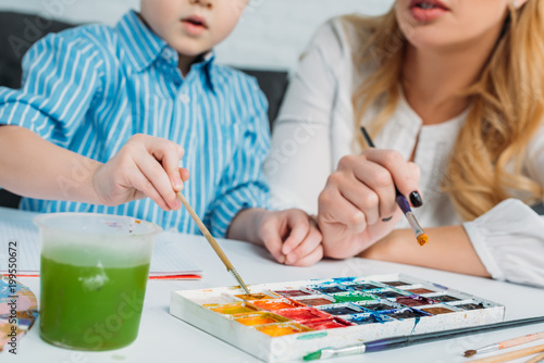 Cropped image of mother and son dipping paintbrushes into paints