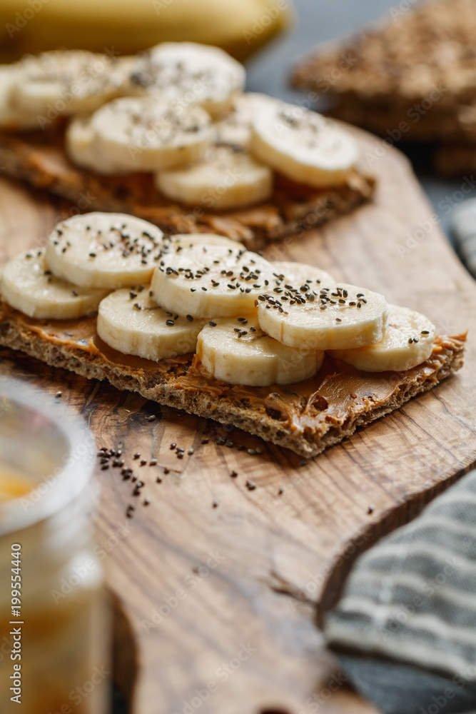 Two multi grain breads with peanut paste, banana slices and chia seeds on a serving wooden board. The concept of healthy fitness breakfast.