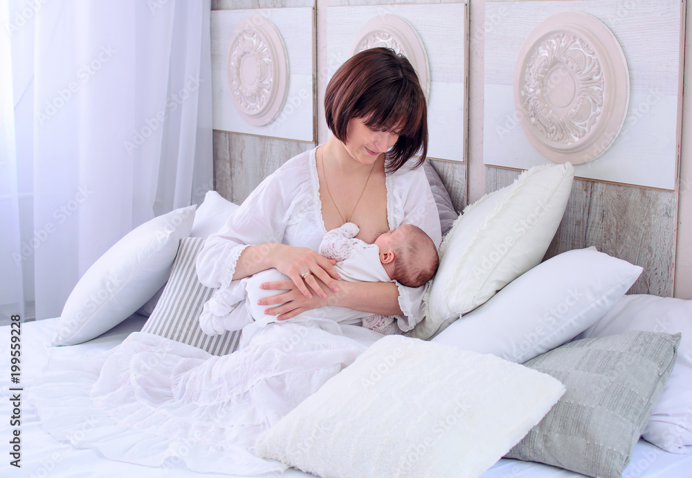 Breastfeeding baby. Young mother holding her newborn child. Mom nursing baby. Woman and new born relax in a white bedroom. Mother breast feeding baby.
