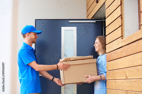 Fotografie Smiling delivery man in blue uniform delivering parcel box to recipient - courier service concept