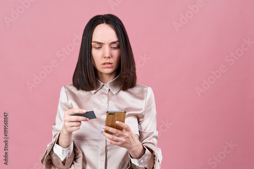 stylish young brunette woman with a serious concentrated expression uses card to pay for purchases in Internet using a smartphone