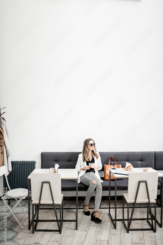 Stylish business woman in white jacket sitting during the coffee break in the modern cafe interior. Wide view with copy space on the wall