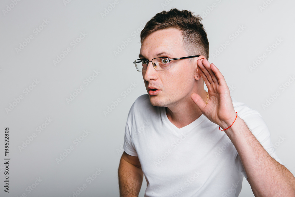 Curious handsome young man in shirt overhearing rumors over grey background. Curious handsome young man in shirt overhearing rumors over grey background.