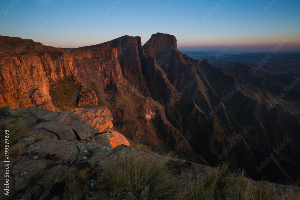 Landscape image of dawn breaking high on top of the Drakensberg Amphitheatre in South Africa. Golden light illuminates cliffs of Sentinel Peak with a clear blue sky as background.