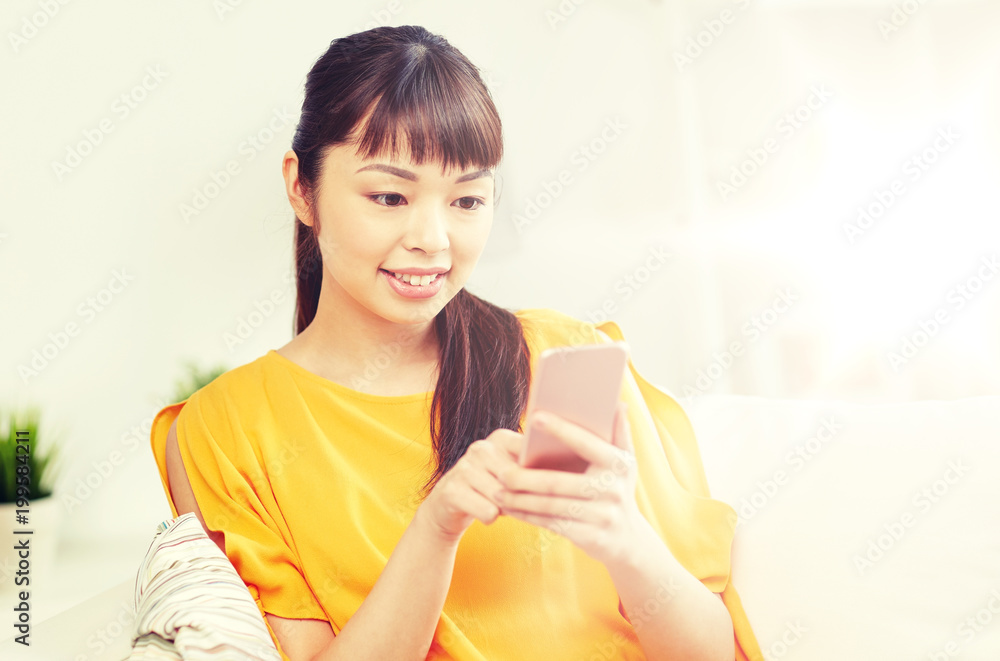 people, technology, communication and leisure concept - happy young asian woman sitting on sofa and texting message on smartphone at home people, technology, communication and leisure concept - happy young asian woman sitting on sofa and texting message on smartphone at home