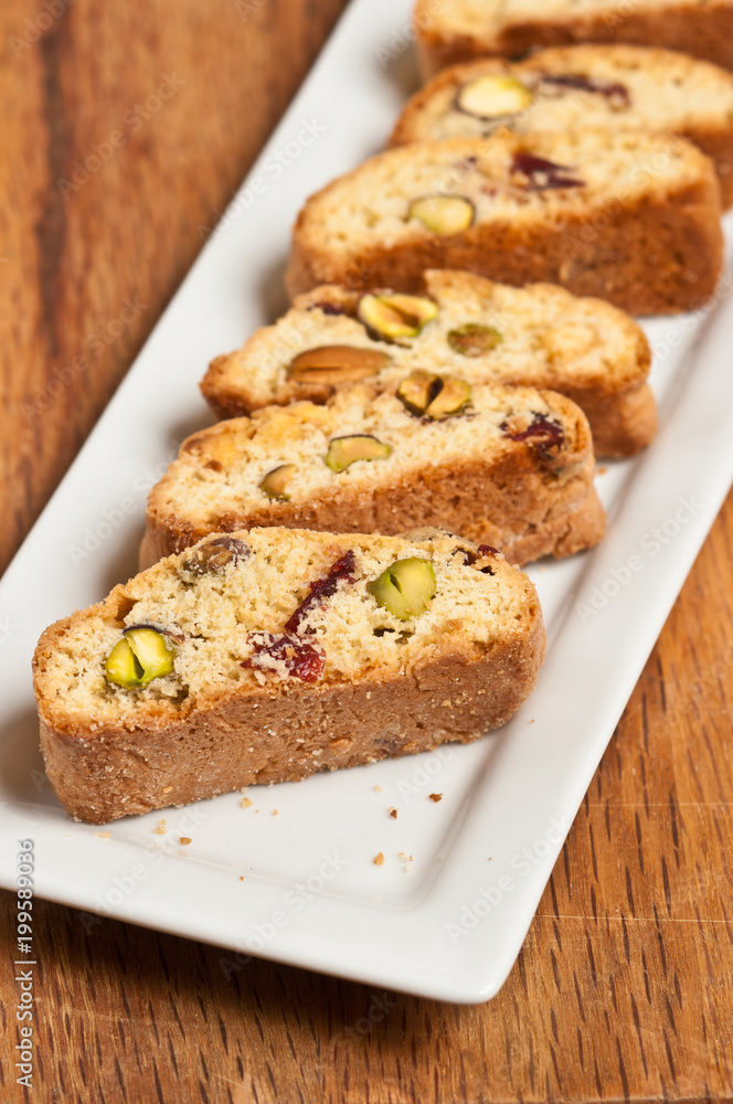 Top view, close up of freshly baked cranberry, pistachio, white chocolate biscotti on a rectangular, white plate on a wood cutting board Top view, close up of freshly baked cranberry, pistachio, white chocolate biscotti on a rectangular, white plate on a wood cutting board
