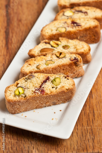 Top view, close up of freshly baked cranberry, pistachio, white chocolate biscotti on a rectangular, white plate on a wood cutting board 