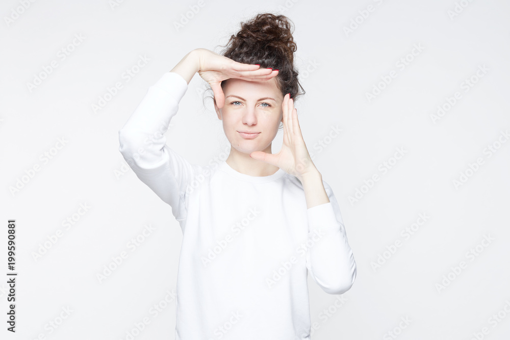Indoor studio shot of young attractive European female with curly hair bun smiling at camera and gesturing finger frame, while standing against isolated white wall background. Body language concept. Indoor studio shot of young attractive European female with curly hair bun smiling at camera and gesturing finger frame, while standing against isolated white wall background. Body language concept.