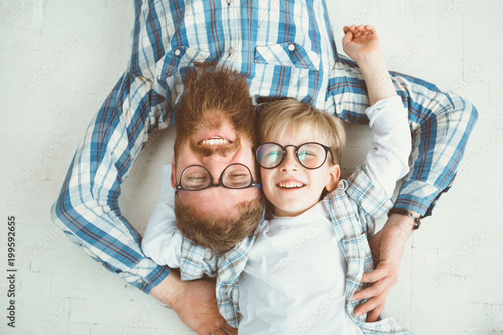 Top view of cute little boy and his handsome young beard dad, both in eyeglasses, smiling while lying with hands behind head on white floor. - obrazy, fototapety, plakaty 