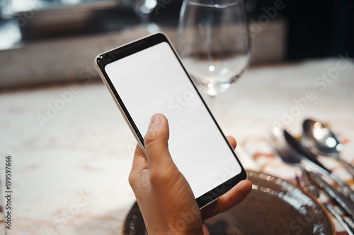 Young brunette girl holding mobile phone while waiting meal. Focus is on hands and mobile