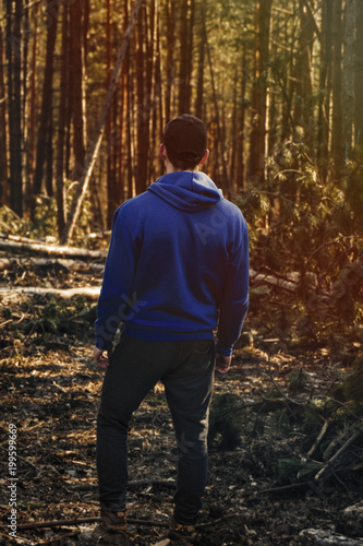 Young caucasian man  hiker  standing in the forest and enjoying perfect view on trees. Man relaxing and chilling in the forest at sunset  image from his back .