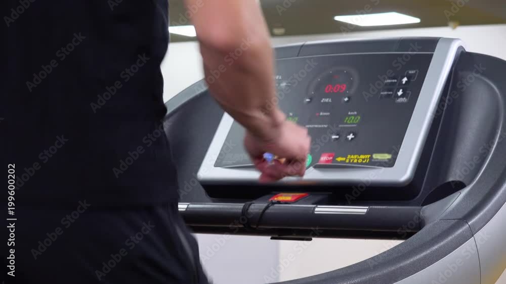 A fit man works on a screen of a treadmill in a gym, then breaks into a jog - closeup from behind A fit man works on a screen of a treadmill in a gym, then breaks into a jog - closeup from behind