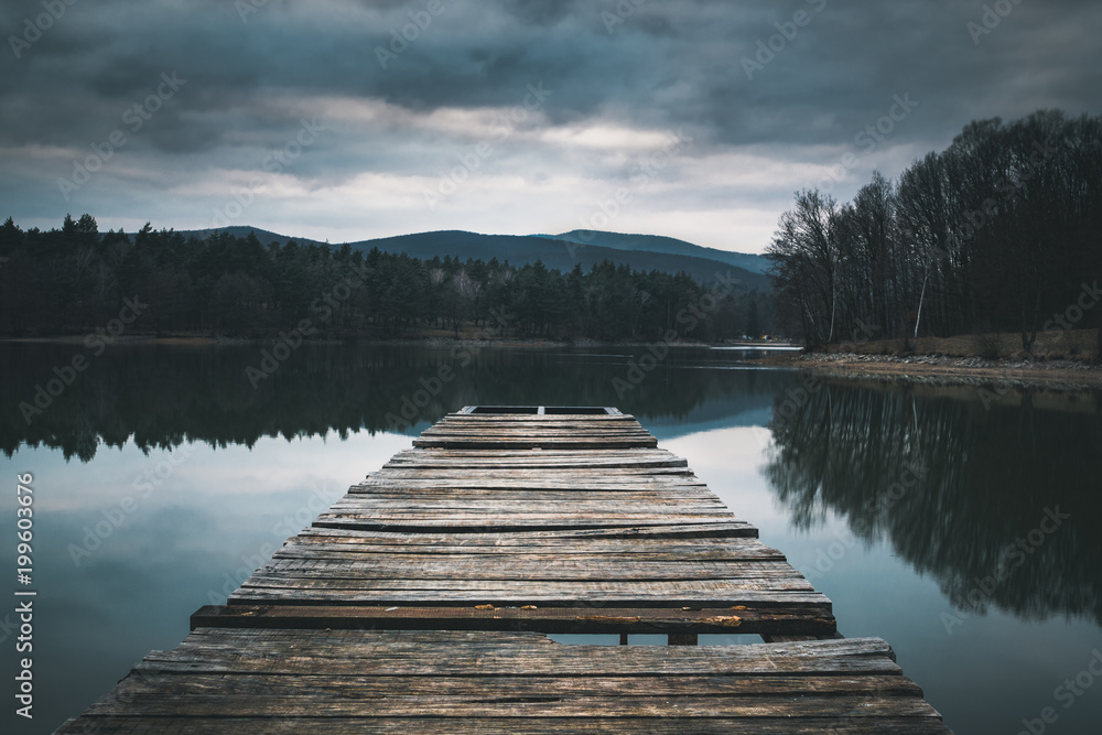Fototapeta premium Mole (pier) on the lake. Wooden bridge in forest in spring time with blue lake. Lake for fishing with pier. Dark and Foggy lake with hills.