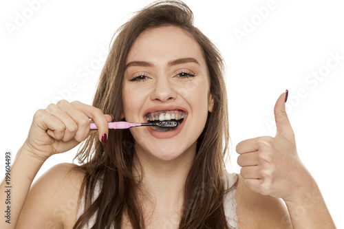 young woman brushing her teeth with a black tooth paste with active charcoal, and black tooth brush on white background, and showing thumbs up photo