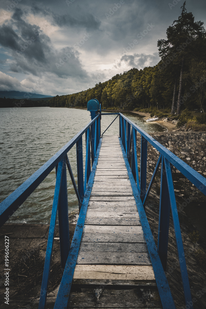 Fototapeta premium Mole (pier) on the scandinavian lake with motorboat on side and misty hills. Blue wooden bridge in forest in autumn time with blue lake. Lake for fishing with pier and powerboat. Foggy lake with hills