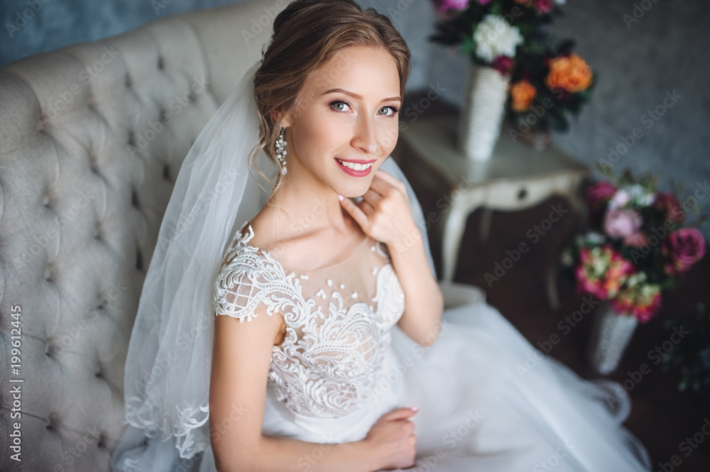 The beautiful bride sits on the couch in a lace dress and smiles sweetly. Sweet bride in a gray studio. Portrait of the bride. The beautiful bride sits on the couch in a lace dress and smiles sweetly. Sweet bride in a gray studio. Portrait of the bride.