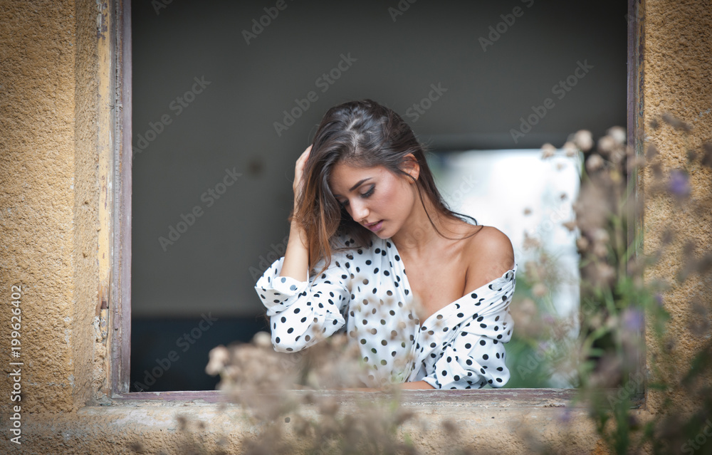 Fototapeta premium the beautiful and sexy girl with long hair stands by the window.Portrait of a pensive woman looking away through a window.Attractive girl with white blouse looks out of the window . 