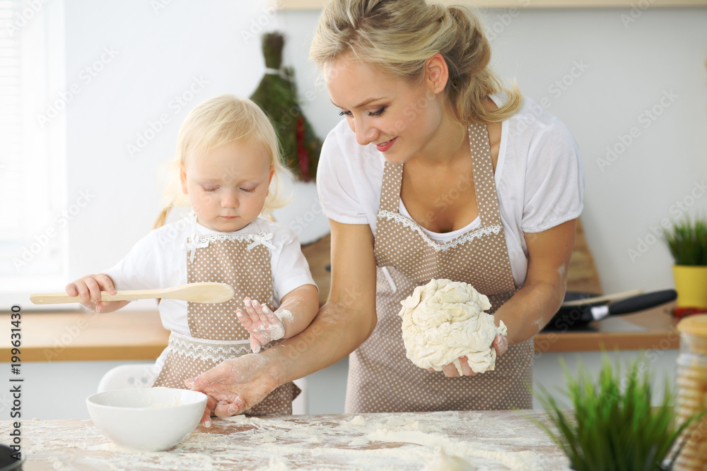 Little girl and her blonde mom in red aprons  playing and laughing while kneading the dough in the kitchen. Homemade pastry for bread, pizza or bake cookies