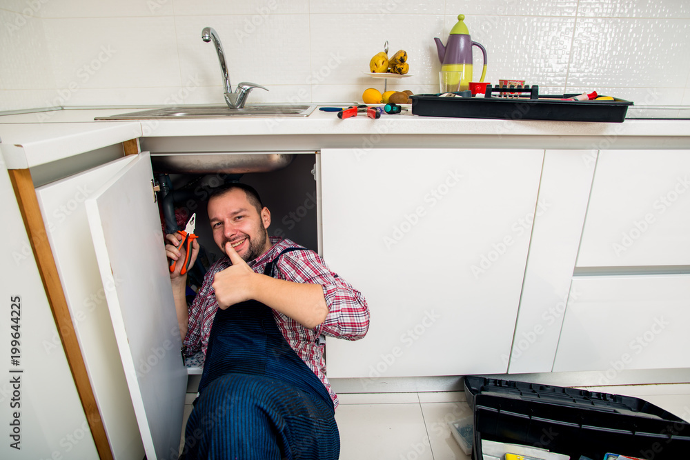 Handyman Lying On Floor Repairing Sink In Kitchen Handyman Lying On Floor Repairing Sink In Kitchen