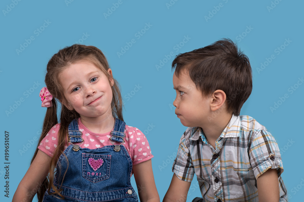 Close up emotional portrait of the smiling of the little girl and angry boy on blue background in studio. Close up emotional portrait of the smiling of the little girl and angry boy on blue background in studio.